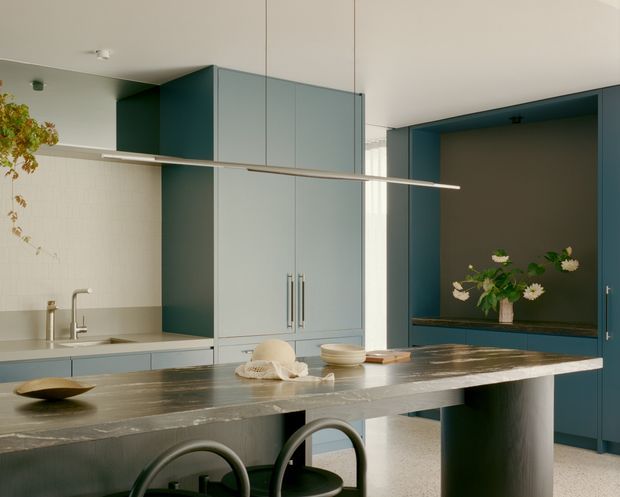 A refined contemporary kitchen with deep teal-blue cabinetry, a pale tiled splashback, and a long dark stone island with integrated seating. Full-height central pantry-style cabinetry divides the room, while a recessed dark-blue display niche with flowers adds depth. A slim linear pendant light spans the island, and soft natural light creates a calm, sophisticated atmosphere.