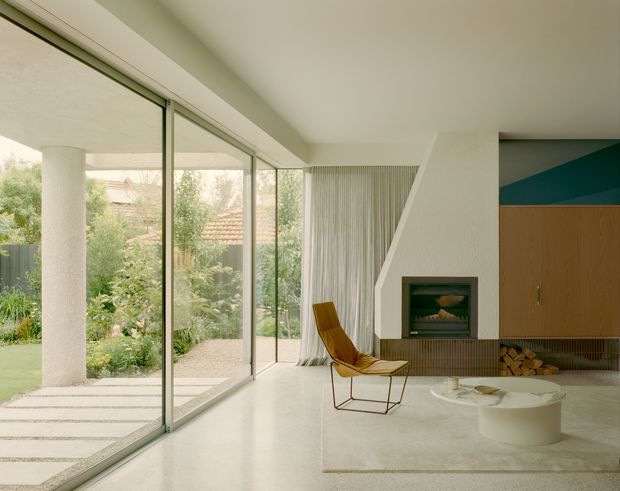 A bright, open-plan mid-century-inspired living room with full-height glazed sliding doors opening to a landscaped garden. The room features a sculptural angled fireplace surround with built-in timber cabinetry and firewood storage, a mustard leather lounge chair, a low round coffee table, polished pale flooring, and sheer vertical curtains. A covered outdoor area and garden are visible through the glazing.