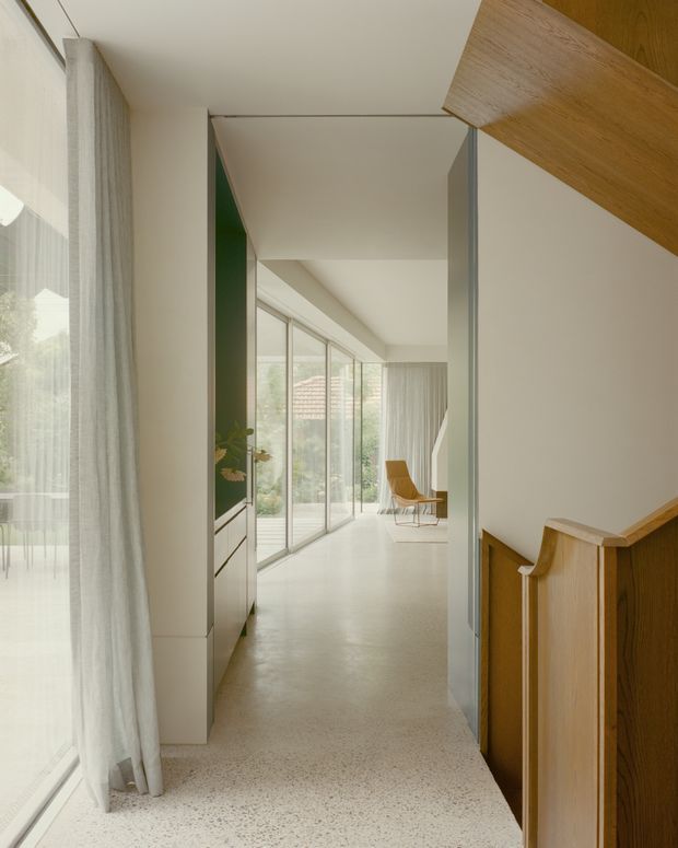 A bright, minimalist residential hallway or landing viewed beside a timber staircase. The space features pale terrazzo flooring, white walls and ceiling, a slim dark green feature wall with built-in cabinetry, full-height glazed doors or windows with sheer curtains, and warm timber stair elements. A small seating area is visible at the far end, with greenery beyond the glazing.