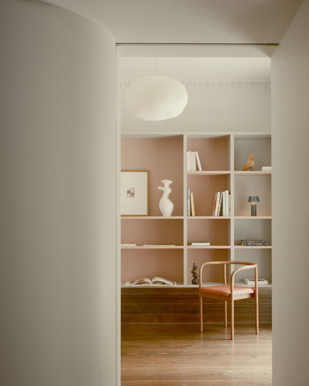 A softly lit, refined home office or study viewed through a wide doorway. The room features a built-in white open shelving unit with warm blush-pink backing, books and restrained decorative objects, a dark timber built-in cabinet beneath, pale timber flooring, and a sculptural upholstered wooden chair. A large rounded pendant light hangs in front of the shelving, while curved plastered wall and ceiling forms frame the opening.