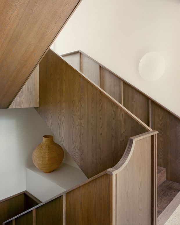 Interior view of a sculptural timber-lined staircase and landing. Warm wood panelling forms the stair balustrades and angled wall surfaces, with a curved handrail detail, pale plaster walls, and a built-in display shelf holding a woven earthen-toned vase. A round wall light adds a subtle focal point.