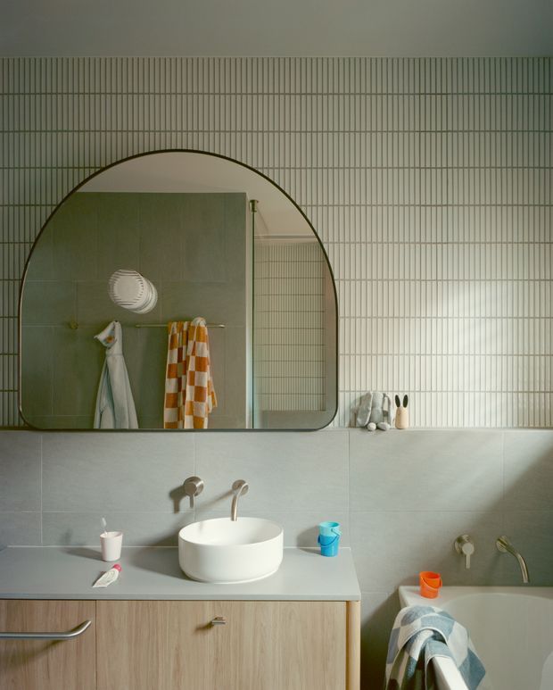 A contemporary bathroom with a pale grey tiled vanity wall, integrated timber-look cabinetry, a white vessel basin, brushed metal wall-mounted tapware, and a partially visible bathtub. A large rounded-arch mirror reflects a tiled shower area with a frameless glass screen and hanging towels. The room uses soft natural light and muted green-grey tones with warm wood accents.