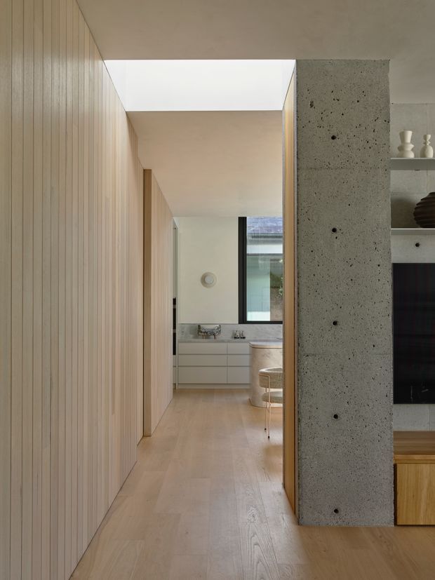 A refined modern interior hallway leading toward a bright living or dining area. The space features extensive light timber vertical wall panelling and matching timber flooring, a raw textured concrete structural column, a linear skylight, pale built-in cabinetry, and partial views of shelving, a television, and upholstered seating. The composition emphasises clean lines, natural materials, and soft neutral tones.