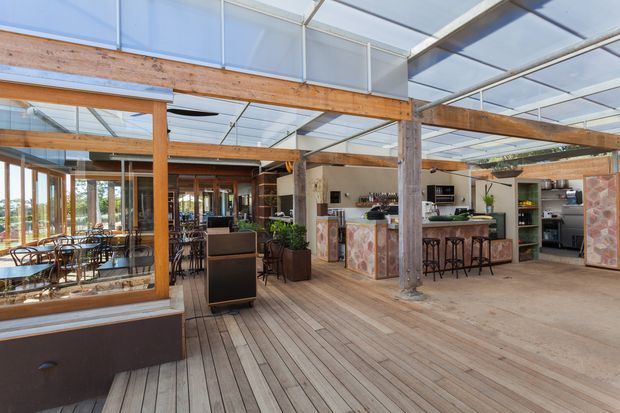A spacious, light-filled commercial hospitality interior, likely a restaurant or café, with extensive glazing and a translucent roof, exposed timber beams and posts, timber decking, dining tables, a central service counter, and a built-in bar with stools. The fit-out combines warm natural timber, textured stone-patterned counter surfaces, and dark metal details.