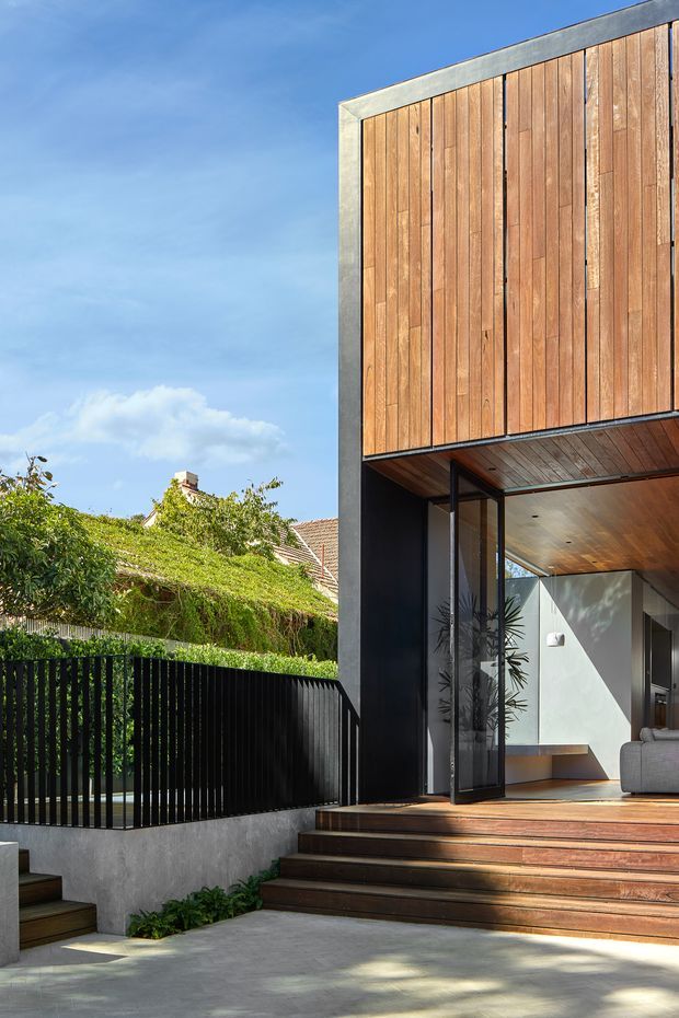 A contemporary multi-level residential facade viewed from the front, featuring a tall timber-clad upper volume framed in concrete or grey metal, dark lower wall surfaces, extensive glazing, and broad timber entry steps. The open doorway reveals part of a minimalist living room with a grey sofa, timber ceiling, and built-in bench. A black vertical-slat fence and landscaped neighbouring residential setting are visible alongside the entrance.