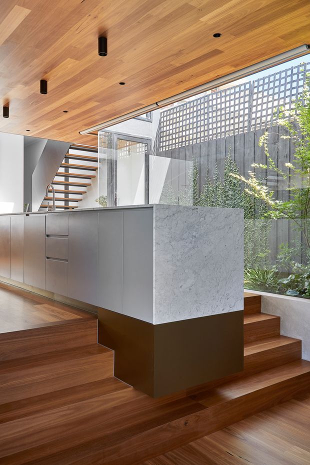 A contemporary open-plan kitchen and stairway interior with a large stone island, matte grey cabinetry, warm timber flooring and a timber-lined ceiling. Floating timber stairs with dark metal supports rise beside full-height glazing, which overlooks a planted courtyard screened by a perforated masonry or concrete wall. Recessed and cylindrical black ceiling lights add contrast to the warm, natural material palette.