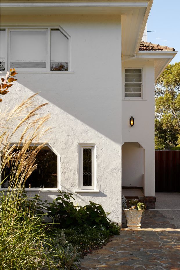 A sunlit exterior view of a stucco-clad residential house, showing white rendered walls, projecting eaves, decorative window trims, leaded-style glazing, a tiled roof edge, and a covered side entry or passage. A flagstone-paved path and landscaped planting occupy the foreground, with strong angular sunlight casting shadows across the facade.