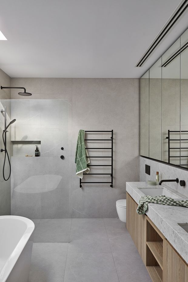 A contemporary bathroom with large light-grey tiles covering the walls and floor. The room includes a freestanding white bathtub in the foreground, a glass-enclosed shower with black fixtures, a wall-mounted black heated towel rail, a toilet, and a timber vanity with a pale stone benchtop and integrated basin. Full-height mirrored cabinetry spans the right wall, reflecting the towel rail and vanity. The design has clean lines, restrained grey and timber tones, and black hardware.
