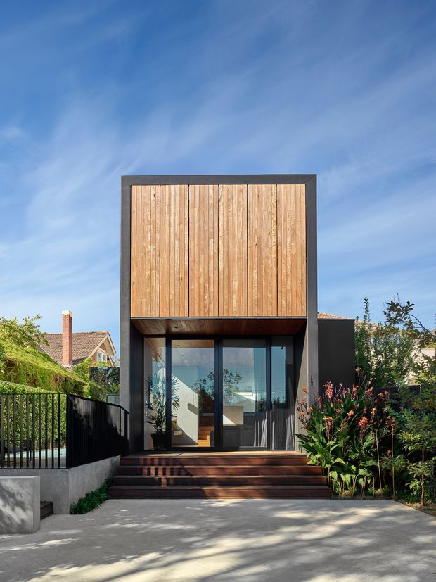 Front exterior view of a compact contemporary two-storey home extension or pavilion. The facade combines a dark charcoal rectangular frame and lower glazing with a prominent upper section clad in vertical warm timber boards. Broad timber steps lead to full-height glazed doors, with a black metal balustrade and landscaped planting around a paved forecourt. A pitched-roof neighbouring house is visible in the background under a bright blue sky.