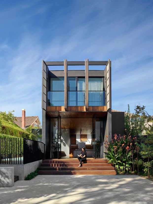Front exterior view of a narrow multi-storey contemporary house with a strongly glazed upper level, dark framed facade, timber-lined balcony and entry soffit, and broad timber steps leading to a recessed entrance. The house is set within a landscaped suburban garden, with neighbouring traditional homes visible to either side.