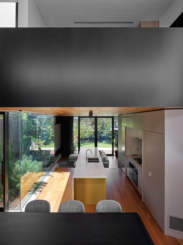 A contemporary open-plan kitchen and dining area viewed from an elevated position. A long white kitchen island with integrated sink and gold-toned end panel sits centrally, with dark cabinetry and integrated appliances along the right wall. Warm timber flooring and a timber-lined ceiling contrast with charcoal walls and joinery. Full-height glazing opens the room to a lush garden, while a dark dining table and upholstered chairs occupy the foreground.