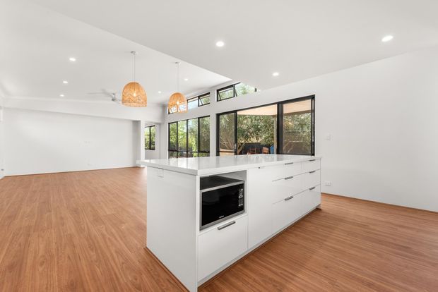 Bright open-plan kitchen and living area with white minimalist cabinetry, a large white island with integrated microwave, warm timber-look flooring, black-framed glazing, and high clerestory windows. Two woven pendant lights and recessed ceiling lights add warmth while the expansive glazing connects the interior to greenery outside.