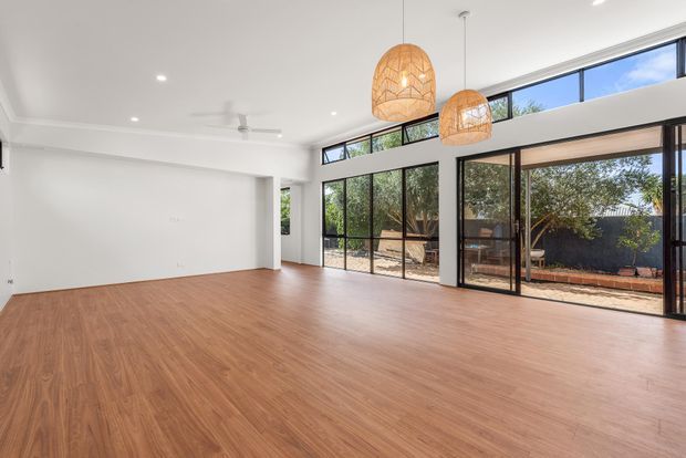 A bright, spacious open-plan interior with warm timber-look flooring, white walls and ceiling, extensive black-framed floor-to-ceiling glazing, and high clerestory windows opening to a landscaped outdoor area. Two woven pendant lights and a ceiling fan add visual interest to the otherwise largely unfurnished room.