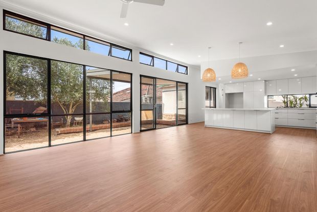 Bright open-plan interior combining a spacious living and dining area with a contemporary white kitchen. The room features extensive black-framed floor-to-ceiling glazing and clerestory windows, warm timber-look flooring, a white ceiling with recessed lighting, and two woven pendant lights over the kitchen island. A courtyard and adjacent brick building are visible outside.