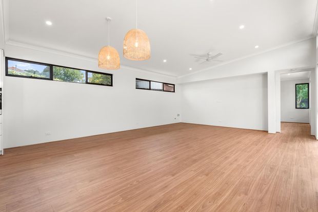 Bright, spacious open-plan interior with white walls and ceiling, warm timber-look flooring, narrow horizontal windows, recessed lighting, woven pendant lights, and a ceiling fan. The room is largely unfurnished, with an opening leading to an adjacent area on the right.