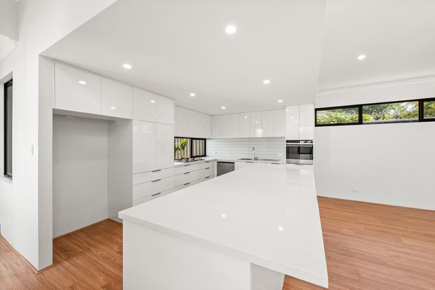 Bright, open-plan contemporary kitchen with extensive glossy white cabinetry, a large white island benchtop, integrated appliances, white tiled splashback, recessed downlights and warm timber flooring. High horizontal windows provide natural light, while a generous open area connects the kitchen to the adjacent living or dining space.