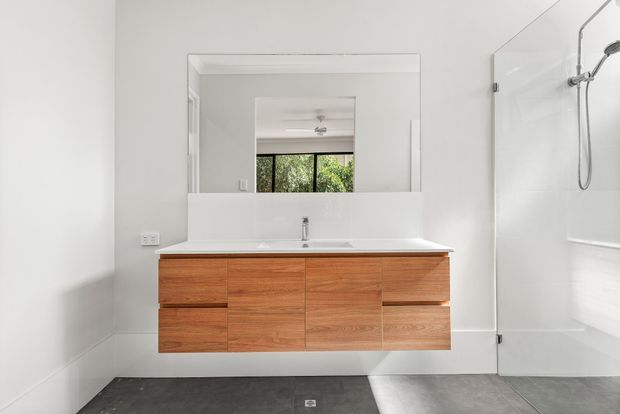 Bright contemporary bathroom featuring a floating warm timber vanity with integrated white basin and broad mirror, alongside a frameless glass shower with chrome fittings. The room has white walls and a dark grey tiled floor, with natural greenery visible through the reflected window.