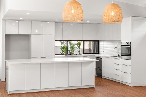 Bright contemporary kitchen with extensive white flat-panel cabinetry, a large central island, white benchtops, integrated appliances, and a window above the sink. Warm timber flooring and two woven pendant lights add natural texture and warmth.