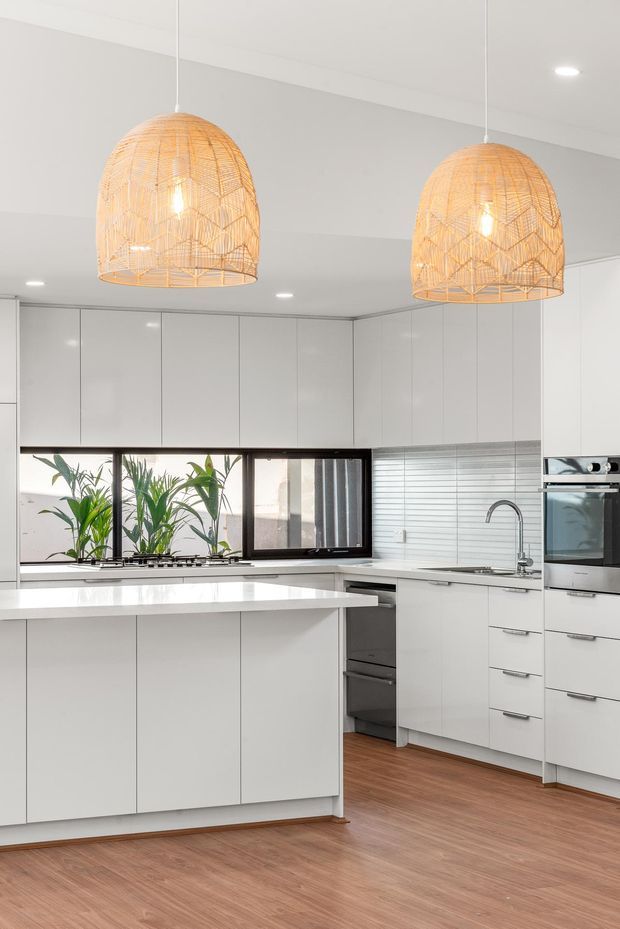 Bright contemporary kitchen with extensive glossy white handleless cabinetry, a large white island, integrated stainless-steel appliances, a white horizontal tile splashback, dark-framed windows, and warm timber-look flooring. Two oversized woven pendant lights add natural texture above the island.