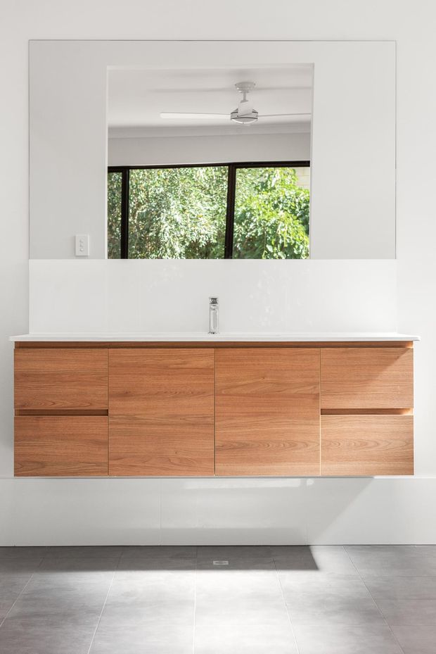 A bright, minimalist bathroom vanity with wall-mounted warm timber cabinetry, a white benchtop and integrated basin, large frameless mirror, and dark-framed window overlooking greenery. Large grey floor tiles and white walls create a clean contemporary interior.