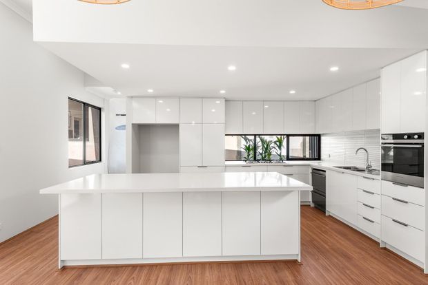 Bright, spacious contemporary white kitchen with extensive glossy handleless cabinetry, a large central island, integrated appliances, white benchtops, timber-look flooring, recessed ceiling lights, and a window above the sink.
