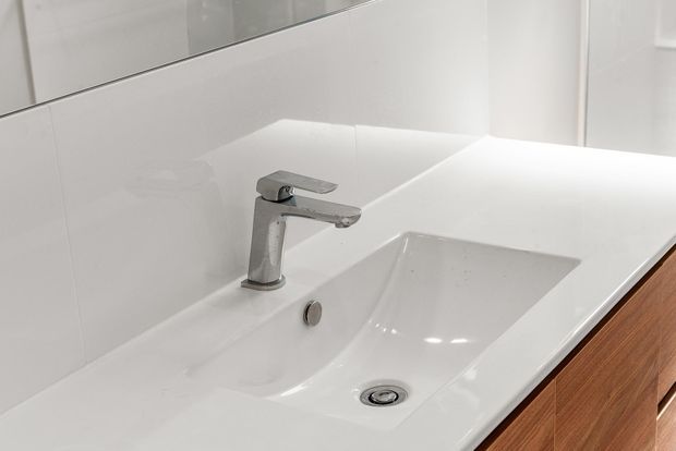 Close-up of a clean, contemporary bathroom vanity with an integrated white rectangular basin and chrome single-lever mixer tap. The vanity has white glossy surfaces and warm timber cabinetry below, with white wall finishes and a mirrored upper section.