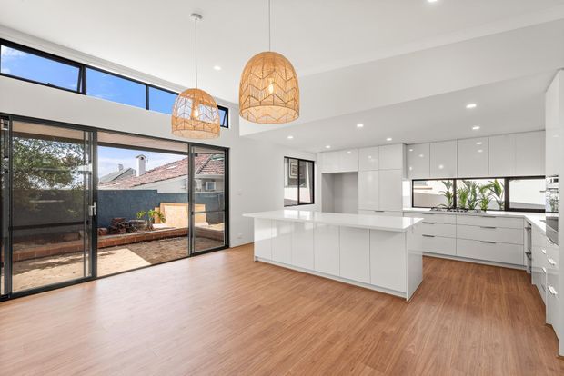Bright open-plan kitchen and living area with a large white island, glossy white cabinetry, integrated appliances, warm timber flooring, and expansive black-framed glazing opening to an outdoor courtyard. Two woven pendant lights add natural texture above the living and dining zone.