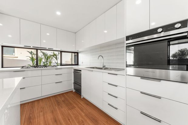 Bright contemporary white kitchen with extensive handleless-style cabinetry, integrated stainless-steel appliances, a horizontal window above the benchtop, a tiled splashback, and warm timber flooring. The compact layout includes a peninsula or work surface and a stainless-steel sink beneath the window.