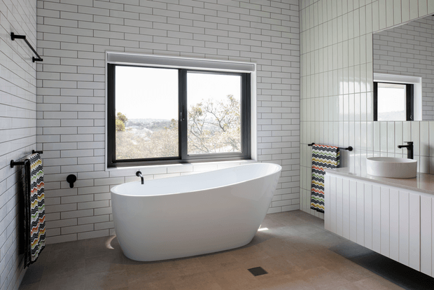 Bright contemporary bathroom featuring a freestanding white bathtub beneath a black-framed window, white horizontal wall tiles, vertical white panelled joinery, a large wall mirror, floating vanity with white basin, and black tapware and towel rails. The room has a warm grey floor and views through the window.