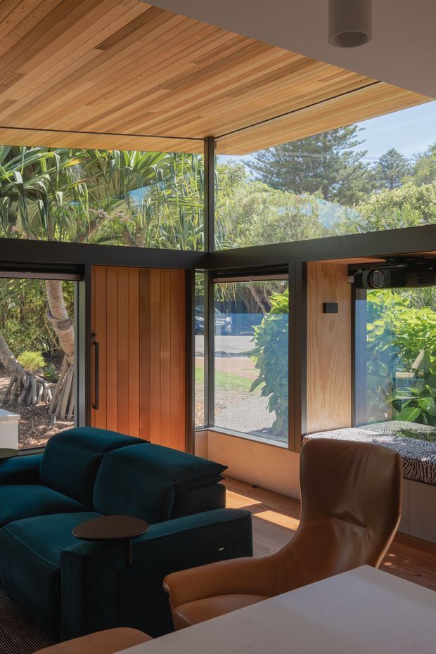 Warm, modern living space with a teal sectional sofa and tan leather lounge chair, framed by extensive floor-to-ceiling glazing and a timber-lined ceiling. A vertical timber entry door and dark metal window framing contrast with the natural wood finishes, while lush planting is visible outside. A large white surface in the foreground may be a kitchen island or table.