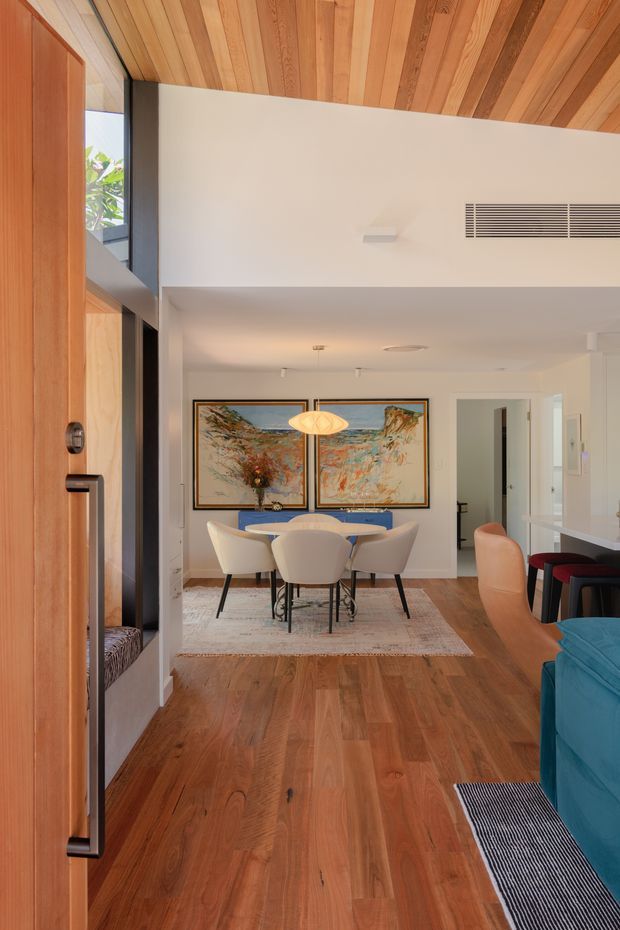 A warm contemporary open-plan interior viewed from the entry, with a timber pivot door and matching timber-lined ceiling. The space features natural timber flooring, a dining area with a round table and upholstered chairs, a large abstract artwork, pendant lighting, and a partial glimpse of a blue sofa and kitchen or bar seating. White walls and built-in air-conditioning keep the room bright and refined.