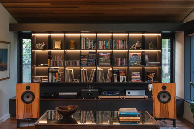 A sophisticated built-in entertainment and library wall dominates the room, with dark shelving illuminated by warm integrated lighting and filled with books, records, and display objects. A central hi-fi and turntable setup sits above dark cabinetry, flanked by large timber-finished speakers. The room also features a reflective glass coffee table, a wood-slat ceiling, pale walls, framed artwork, and windows on either side.