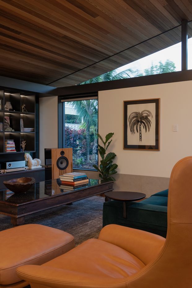 A warm, richly layered living room with a low timber-lined ceiling, dark built-in shelving, large windows opening toward a leafy garden, and a framed botanical artwork. The room includes a dark wood and glass coffee table, hi-fi equipment and speaker, a deep green upholstered chair, and prominent cognac leather lounge seating in the foreground. The palette is earthy and moody, with a refined mid-century character.
