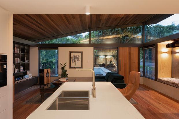 A warm, open-plan contemporary kitchen and living area viewed from behind a large white island bench. The kitchen features an inset stainless-steel sink and minimalist curved tap, dark built-in cabinetry, timber flooring, and a richly timber-lined ceiling. Beyond the island is a compact living area with a dark sofa, leather lounge chair, shelving, artwork, and indoor plants. Extensive glazing and clerestory windows bring in views of lush vegetation, while a timber entry door and built-in window seat add warmth.