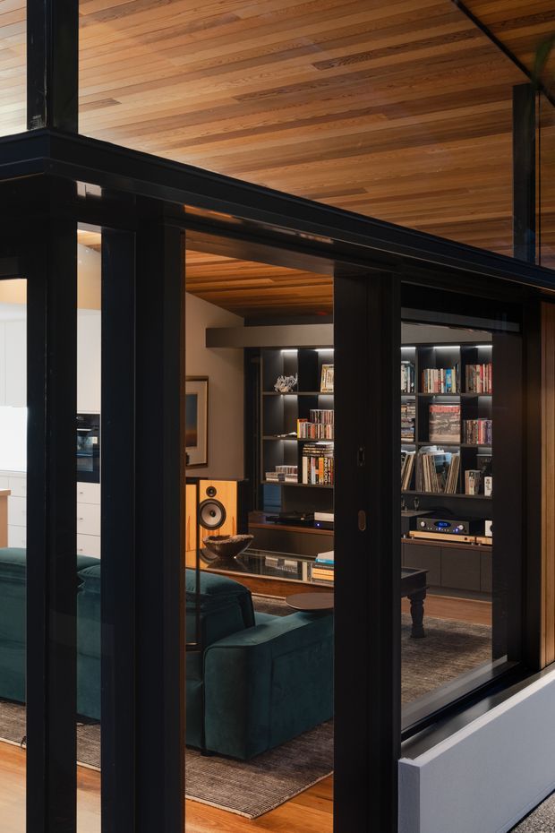 Interior view through black-framed glazed doors or windows into a refined media and living room. The room features a warm timber-lined ceiling, dark built-in shelving filled with books and records, integrated audio equipment, large speakers, a deep teal sofa, a coffee table, and a rug. The prominent black framing and warm wood surfaces create a sophisticated, cozy atmosphere.