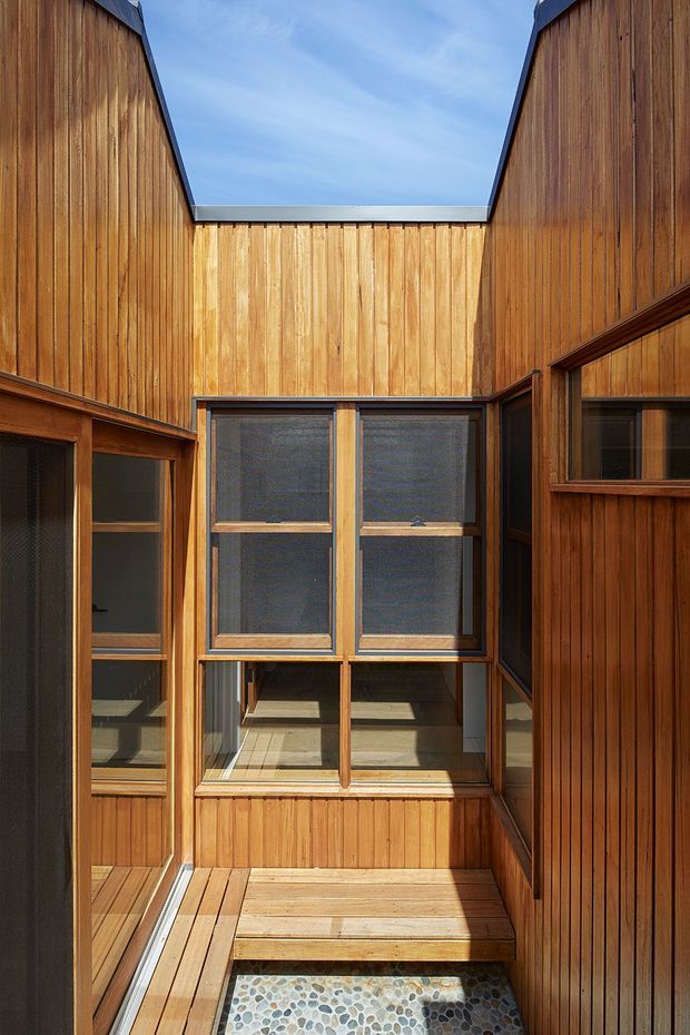 A compact open-air courtyard or lightwell enclosed by tall vertical timber-clad walls. Timber-framed glazed openings surround a small stepped timber deck, with a pebble-finished lower area visible at the base. Bright daylight enters through the open roof, creating a warm, secluded architectural space with strong geometric lines.