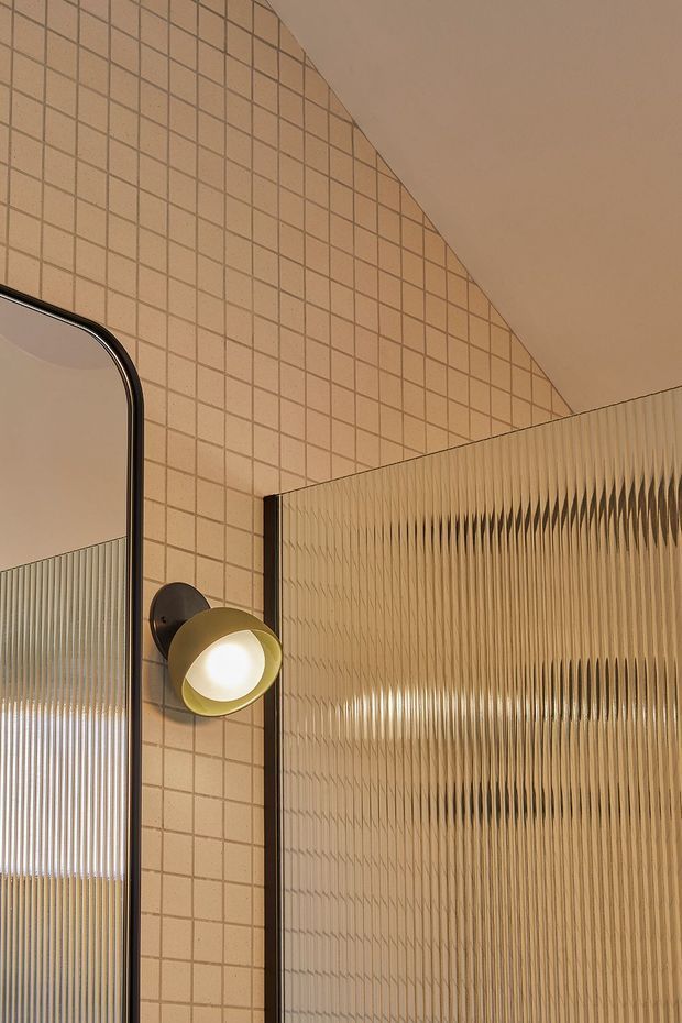 Close-up interior detail featuring a warm beige tiled wall with a fine square grid pattern, a rounded rectangular mirror with a dark metal frame, a sculptural wall-mounted globe light, and a vertically ribbed translucent glass partition. The composition is warm, tactile, and softly illuminated, with strong geometric and fluted textures.