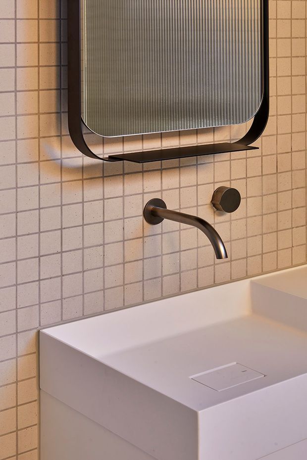 Close-up view of a contemporary bathroom vanity with a wall-mounted curved bronze tap, matching round control, integrated white basin, and a rounded rectangular mirror with a dark frame. The wall is finished in small square blush-beige tiles with dark grout, creating a warm, refined palette.