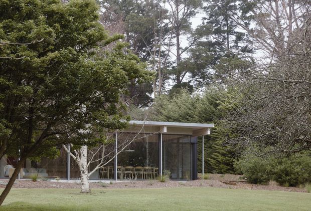 A low, modern pavilion-like building set within a mature landscaped garden. The structure features a broad flat roof with a projecting eave, extensive floor-to-ceiling glazing, slender dark window frames, and pale solid wall panels. Outdoor dining furniture is visible through the glass, while large trees and lawn surround the building.