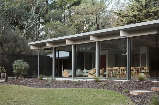 A low-slung modern single-storey house set within a wooded landscape. The building has a broad flat roof with exposed projecting beams, extensive floor-to-ceiling glazing in dark frames, and a minimalist open-plan interior visible through the windows. A landscaped garden with lawn, gravel and native planting surrounds the facade.