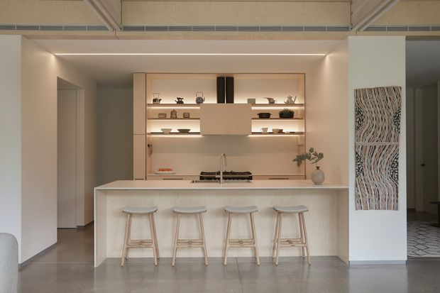 A warm, contemporary open-plan kitchen with a pale stone island and four light timber bar stools. The kitchen features recessed, illuminated open shelving, integrated cabinetry, a compact cooktop and sink, and a softly lit neutral palette. Polished concrete flooring, exposed ceiling services, and a decorative textured wall panel add subtle character. A glimpse of an adjacent room is visible at right.