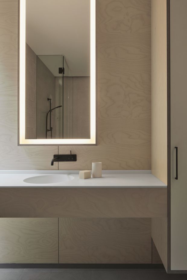 A minimalist bathroom vanity with extensive pale plywood cabinetry and wall panelling, a white integrated basin and countertop, matte black wall-mounted tapware, and a rectangular backlit mirror. A glass shower enclosure is reflected in the mirror, while a tall storage cabinet occupies the right side. The restrained palette and clean, handle-free joinery create a calm contemporary interior.