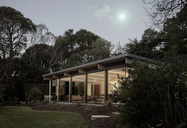 A low-slung, single-storey modern pavilion set within a landscaped garden. The building has a broad flat roof with deep overhangs, slim dark structural posts, extensive floor-to-ceiling glazing, and warm interior lighting visible through the windows. Mature trees and planted borders surround the home, with a lawn and stepping-stone path in the foreground. The image has a subdued, evening atmosphere.