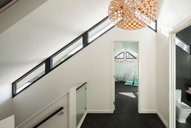 Bright upper-level hallway and stair landing with a steeply sloped ceiling, a long row of black-framed clerestory windows, white walls, dark carpet, and a decorative woven pendant light. The landing opens toward a playful children's bedroom, while a bathroom is visible through a doorway on the right.