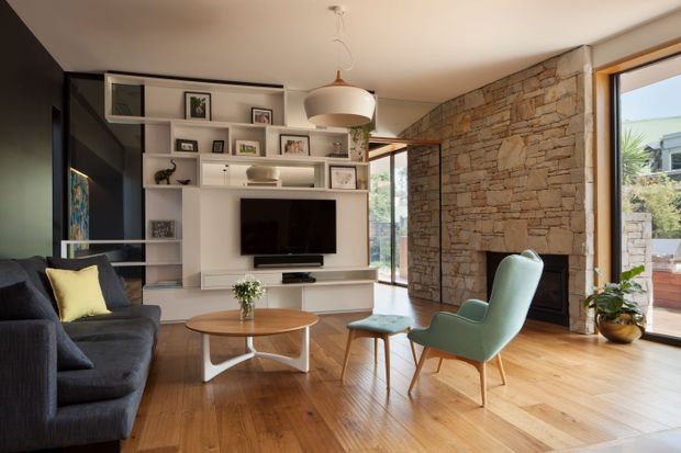 A warm, refined living room with a dark sectional sofa, pale timber flooring, built-in white media and display cabinetry, a sculptural pendant light, and mid-century-inspired lounge chairs. A substantial exposed stone feature wall incorporates a fireplace, while large glazed openings bring in natural light and views to the exterior.