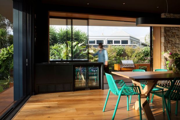 A contemporary indoor-outdoor dining and entertaining space with warm timber flooring, dark ceiling and window framing, large glazed openings, a timber dining table, and bright turquoise chairs. A compact drinks refrigerator and dark built-in cabinetry sit against the glazed wall, while a stone-clad wall and lush planted garden are visible around the perimeter. The space opens directly to an exterior deck or garden and receives abundant natural light.