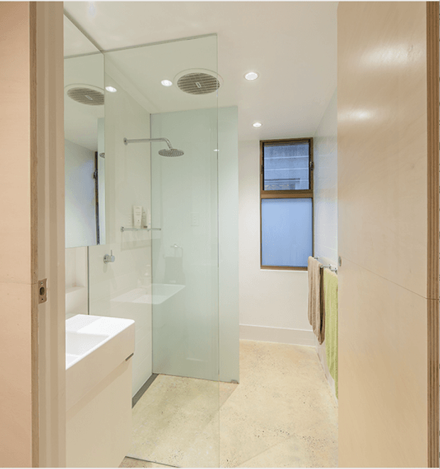 Bright contemporary bathroom viewed through an open doorway, featuring a frameless glass walk-in shower with dual rainfall showerheads, a white floating vanity, large wall mirror, recessed ceiling lights, pale tiled walls, and a small frosted window. The room has a clean, light neutral palette with timber-toned door and wall elements.