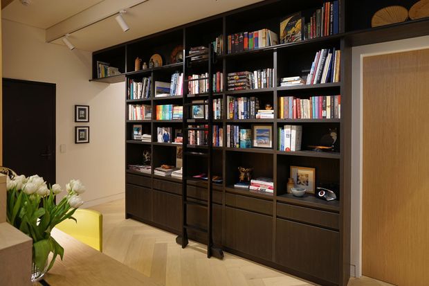 A warm, refined interior centered on a full-height dark timber built-in library wall with extensive open shelving, drawers, books, and decorative objects. A rolling library ladder runs along the shelving. The room includes pale herringbone timber flooring, track lighting, framed artwork, a wood-panelled door, and a partial dining or sitting area with a bright yellow chair and flowers in the foreground.