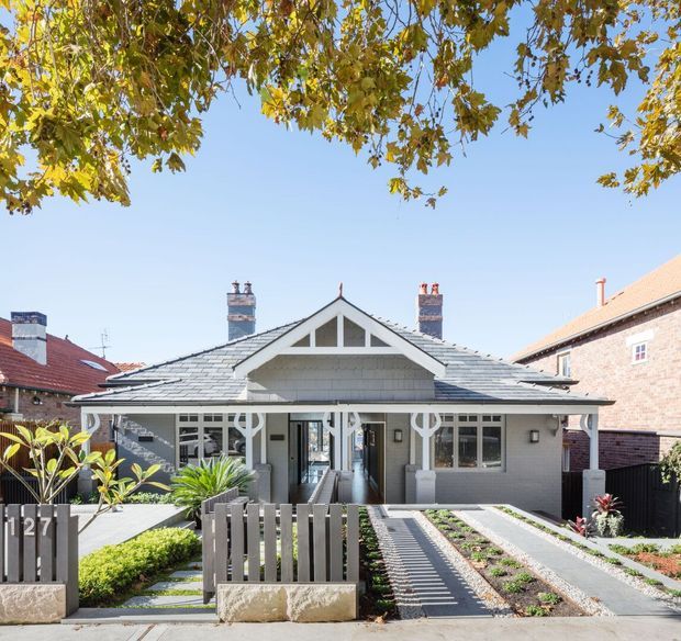 Front exterior view of a single-storey traditional bungalow with a symmetrical gabled roof, prominent front porch, decorative gable detailing, brick chimney stacks, and grey-painted masonry walls. A landscaped front garden includes raised planting beds, paved paths, and a low timber-and-stone boundary fence. Neighbouring homes are visible on either side beneath a clear sky.
