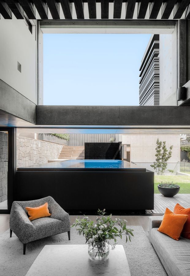 A contemporary living room opens directly onto a courtyard with a raised swimming pool and stepped landscaping. The interior features a grey upholstered armchair and sofa with vivid orange cushions, a pale rug, a white marble-look coffee table, and a large black built-in architectural volume. Strong black timber ceiling beams, concrete surfaces, expansive glazing, and a framed double-height opening create a dramatic indoor-outdoor composition.